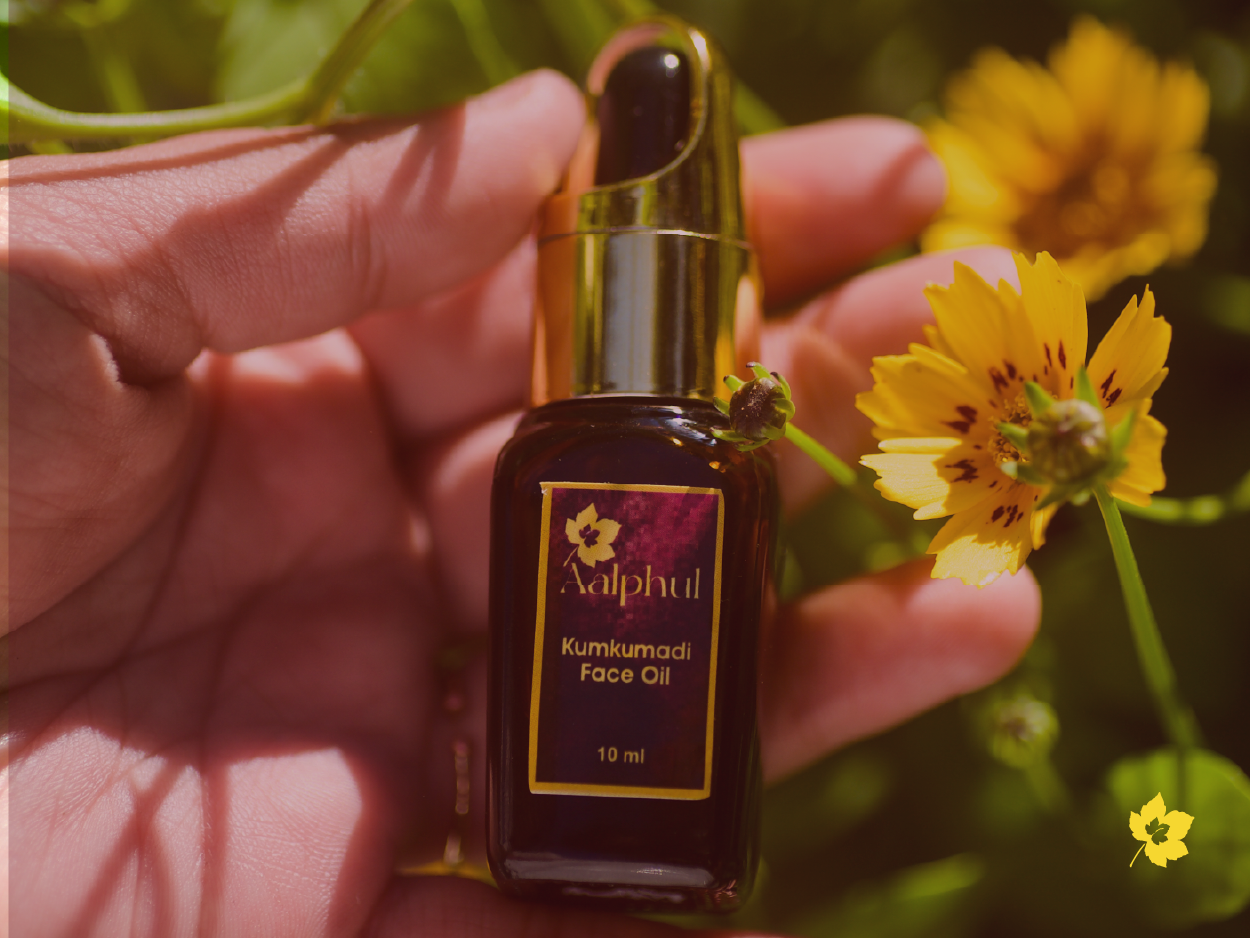 Hand holding a bottle of Aalphul Kumkumadi Face Oil with yellow flowers in the background